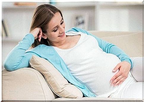 A pregnant woman is holding her stomach and sitting on a couch, smiling.